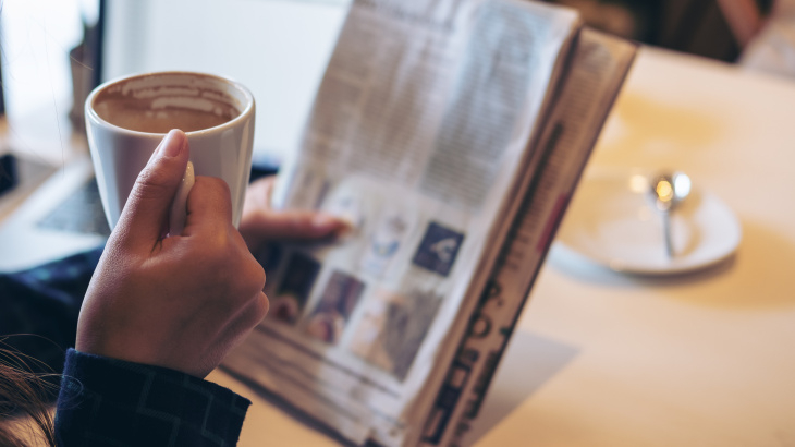 Eine Person liest Zeitung und hält dabei eine Tasse mit einem Heißgetränk.